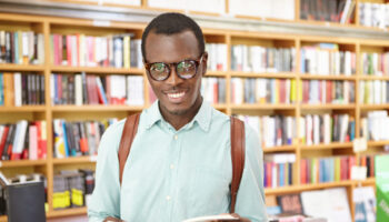 cheerful-young-fashionable-black-man-wearing-glasses-standing-library-with-shelves-books